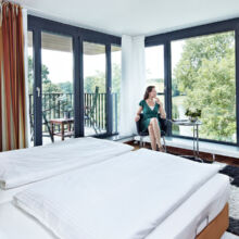 Eine Frau sitzt an einem kleinen runden Tisch neben großen, bodentiefen Fenstern in einem hellen, modernen Hotelzimmer mit einem Doppelbett, gestreiften Vorhängen und Blick auf grüne Bäume draußen.