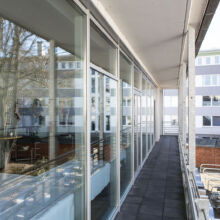 Auf einem langen Balkon mit Glaswänden und Schiebetüren spiegeln sich Bäume und nahe gelegene Gebäude; darunter sind Fahrräder geparkt, und durch die Fenster ist ein Innenhof zu sehen.