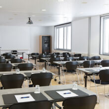 Ein helles, modernes Klassenzimmer mit Reihen von Tischen und schwarzen Stühlen, jeder Tisch mit einem Notizblock, einem Stift und einem Glas Wasser ausgestattet. Große Fenster säumen eine Wand, und von der Decke hängt ein Projektor.