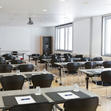 Ein helles, modernes Klassenzimmer mit Reihen von Tischen und schwarzen Stühlen, jeder Tisch mit einem Notizblock, einem Stift und einem Glas Wasser ausgestattet. Große Fenster säumen eine Wand, und von der Decke hängt ein Projektor.