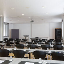 Ein moderner Konferenzraum mit Reihen schwarzer Stühle und Tische, die mit Notizblöcken, Stiften und Wassergläsern ausgestattet sind und auf eine weiße Projektionsfläche im Vordergrund blicken. Große Fenster säumen eine Wand und lassen natürliches Licht herein.