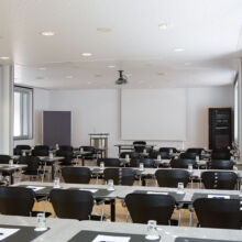 Ein moderner Konferenzraum mit Reihen schwarzer Stühle und Tische, die mit Notizblöcken, Stiften und Wassergläsern ausgestattet sind und auf eine weiße Projektionsfläche im Vordergrund blicken. Große Fenster säumen eine Wand und lassen natürliches Licht herein.