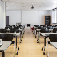 Ein modernes, leeres Klassenzimmer mit Reihen von schwarzen Stühlen und Tischen, jeder mit einem Glas Wasser und einem Notizblock. Große Fenster säumen die rechte Wand, und an der Decke ist ein Projektor angebracht, der auf ein Whiteboard gerichtet ist.