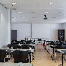 Ein moderner, leerer Konferenzraum mit schwarzen, in Reihen angeordneten Stühlen und Tischen, einem Podium an der Stirnseite, einem Projektor an der Decke und großen Fenstern, die Tageslicht hereinlassen.