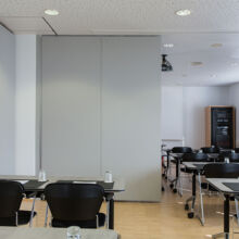 Ein modernes Klassenzimmer mit schwarzen Stühlen und grauen Tischen, die in Reihen angeordnet sind. Eine große graue Trennwand unterteilt den Raum, und durch die Fenster auf der rechten Seite fällt Sonnenlicht ein. In der hinteren Ecke ist ein Schrank zu sehen.