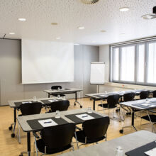 Ein moderner Konferenzraum mit Reihen von Tischen und schwarzen Stühlen, jeder Tisch mit Notizblöcken, Stiften und Wassergläsern ausgestattet. Es gibt einen Projektor, eine Leinwand, ein Flipchart und große Fenster, die natürliches Licht hereinlassen.