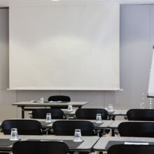 Ein moderner Konferenzraum mit Reihen schwarzer Stühle und Tische, die jeweils mit einem Notizblock, einem Stift und einem umgedrehten Glas ausgestattet sind. An der Stirnseite befinden sich eine leere Projektionsfläche und ein Flipchart mit leerem Papier.