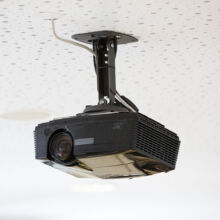 Ein schwarzer, an der Decke montierter Projektor ist mit einer Metallhalterung und einem sichtbaren Kabel an einer weißen, perforierten Decke befestigt.