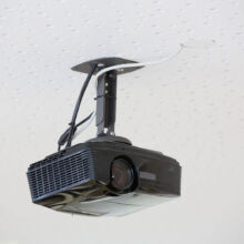 Ein schwarzer Projektor ist an einer weißen, perforierten Decke montiert, wobei die Kabel sichtbar sind, die mit dem Gerät und durch die Decke verbunden sind. Der Projektor ist so positioniert, dass er über Kopf angezeigt werden kann.