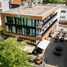 default Hotel SeeZeit am Aasee in Münster. Ein modernes, dreistöckiges Gebäude mit großen Fenstern und einem Schild auf dem Dach mit der Aufschrift "SeeZeit". Im Erdgeschoss sind Sitzgelegenheiten im Freien und Sonnenschirme für die Terrasse aufgestellt. Bäume und benachbarte Gebäude sind in der Nähe zu sehen.