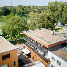 default Luftaufnahme von modernen Wohngebäuden mit Flachdächern, umgeben von Bäumen und einem nahe gelegenen See an einem sonnigen Tag. Einige Balkone sind mit Gartenmöbeln ausgestattet, und Schatten fällt auf die Wege darunter.