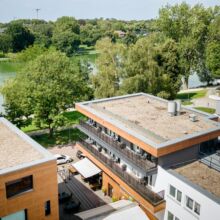 default Luftaufnahme von modernen Wohngebäuden mit Flachdächern, umgeben von Bäumen und einem nahe gelegenen See an einem sonnigen Tag. Einige Balkone sind mit Gartenmöbeln ausgestattet, und Schatten fällt auf die Wege darunter.
