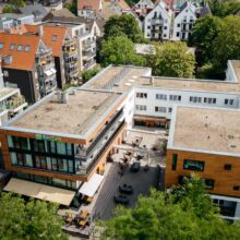 default Luftaufnahme eines modernen, mehrstöckigen Gebäudekomplexes mit Flachdächern, Sitzgelegenheiten im Freien und umliegenden Bäumen. In der Nähe befinden sich Wohngebäude mit Schrägdächern und Grünanlagen.