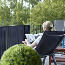 Eine Person mit blondem Haar sitzt auf einem Balkonstuhl und liest eine Zeitung. Neben ihr stehen ein Tisch mit einer Wasserflasche, eine Topfpflanze im Vordergrund und grüne Bäume im Hintergrund.