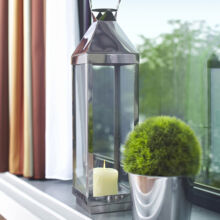 Eine Metalllaterne mit einer weißen Kerze darin und eine kleine grüne Topfpflanze stehen auf einer Fensterbank neben einem großen Fenster mit Vorhängen, aus dem man Bäume sehen kann.