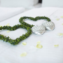 Zwei grüne herzförmige Kränze und zwei silberne Herzornamente liegen auf einer weißen Tagesdecke, umgeben von verstreuten weißen und hellgelben Blütenblättern.