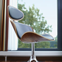 Ein moderner Loungesessel mit geschwungenen Holzpaneelen, schwarzen Lederkissen und einem verchromten Gestell steht auf einem Holzfußboden neben einem großen Fenster mit Bäumen im Freien im Hintergrund.