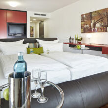 Modernes Hotelzimmer mit einem Doppelbett, roten Schränken, Wandkunst, einem Tablett mit einer Wasserflasche und Gläsern und einem schlichten Dekor. Das Zimmer hat einen Holzfußboden und ein minimalistisches, stilvolles Design.