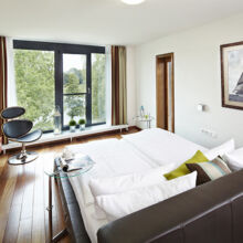 Ein modernes, helles Schlafzimmer mit einem großen Bett, einem gläsernen Beistelltisch, einem eleganten schwarzen Stuhl, Holzfußboden und einem großen Fenster mit Blick ins Grüne. Ein gerahmtes Segelbootbild hängt an der weißen Wand.