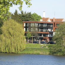 Hinter einem Park mit grünen Bäumen und Menschen, die sich an einem sonnigen Tag an einem Fluss entspannen, steht ein modernes Backsteinwohnhaus mit großen Balkonen.