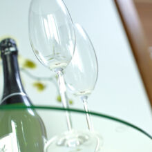 Zwei leere Weingläser stehen neben einer Weinflasche auf einem Glastisch, der Hintergrund ist unscharf.