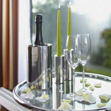 Ein runder Metalltisch mit einer Flasche Champagner in einem silbernen Kühler, zwei leeren Weingläsern, zwei grünen Kerzen in Haltern und weißen Rosenblättern, die auf der Oberfläche verstreut sind. Im Hintergrund sind ein Fenster und Vorhänge zu sehen.