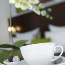 Nahaufnahme einer weißen Teetasse mit Untertasse auf einem Glastisch, daneben ein Teller mit verschiedenen Pralinen und im Hintergrund eine verschwommene weiße Orchideenblüte. Die Umgebung wirkt elegant und modern.