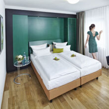 Eine Frau in einem grünen Kleid steht am Fenster eines modernen Hotelzimmers mit zwei sauber gemachten Betten, Holzboden, grüner Akzentwand, einem kleinen Tisch und einem schwarzen Sessel.