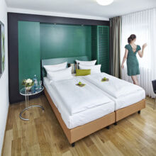 Eine Frau in einem grünen Kleid steht am Fenster eines modernen Hotelzimmers mit zwei sauber gemachten Betten, Holzboden, grüner Akzentwand, einem kleinen Tisch und einem schwarzen Sessel.