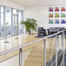 Eine moderne Lobby mit Holzböden, Glasgeländern, hohen Fenstern, Cocktailtischen mit Wasserflaschen, einer Sitzgruppe aus schwarzem Leder, einer großen Topfpflanze und bunten Kunstwerken an der weißen Wand.