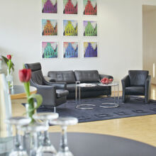 Moderner Lounge-Bereich mit schwarzen Ledersofas und -sesseln auf einem dunklen Teppich, Glastischen mit Wasserflaschen und Gläsern, einer einzelnen roten Tulpe in einer Vase und bunten Kunstwerken an der weißen Wand im Hintergrund.