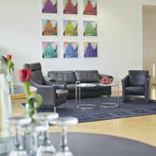 Moderner Lounge-Bereich mit schwarzen Ledersofas und -sesseln auf einem dunklen Teppich, Glastischen mit Wasserflaschen und Gläsern, einer einzelnen roten Tulpe in einer Vase und bunten Kunstwerken an der weißen Wand im Hintergrund.