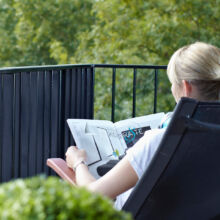 Eine Person mit blondem Haar sitzt auf einem Balkonstuhl und liest eine Zeitung. Eine Flasche Wasser steht auf einem kleinen Tisch neben ihr. Im Hintergrund sind Bäume und Grünpflanzen zu sehen.