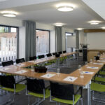 Ein moderner Konferenzraum mit U-förmigen Tischen, schwarzen und grünen Stühlen, Notizblöcken, Stiften und Wassergläsern an jedem Platz; große Fenster und neutrales Dekor schaffen eine helle, professionelle Atmosphäre.