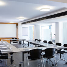 Ein moderner Konferenzraum mit U-förmigen Tischen, schwarzen Stühlen, Notizblöcken und Gläsern, die für eine Besprechung vorbereitet sind. Es gibt eine große Projektionsfläche, ein Flipchart und große Fenster, die natürliches Licht hereinlassen.