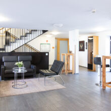 Eine moderne Lobby mit schwarzen Ledersesseln, Stehtischen aus Holz mit Barhockern, einer Treppe mit schwarzem Geländer und hellem Tageslicht. Ein kleiner Teppich und Pflanzen tragen zur einladenden Atmosphäre bei.