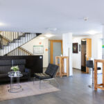 Eine moderne, helle Lobby mit schwarzen Ledersesseln, einem Sofa, Glastischen mit Pflanzen, hohen Holztischen und einer Treppe mit schwarzem Geländer im Hintergrund. Weiße Wände und große Fenster schaffen ein offenes Ambiente.