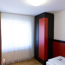 Ein helles, modernes Hotelzimmer mit einem großen Fenster, das von weißen Vorhängen verdeckt wird, einem rot-schwarzen Kleiderschrank, einem roten Schreibtisch mit Lampe und Fernseher und einem Einzelbett mit weißer Bettwäsche.