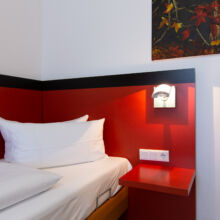 Ein ordentlich gemachtes Einzelbett mit weißer Bettwäsche steht vor einer rot-schwarzen Wand. Eine Wandlampe und ein kleines rotes Regal mit Steckdosen stehen neben dem Bett, und über dem Bett ist ein Teil eines Blumengemäldes zu sehen.