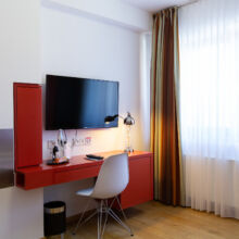 In einem modernen Hotelzimmer stehen ein roter Schreibtisch mit einem darüber montierten Fernseher, ein weißer Stuhl, eine Lampe, eine Flasche Wasser, Gläser und ein Fenster mit durchsichtigen und braunen Vorhängen, die natürliches Licht hereinlassen.