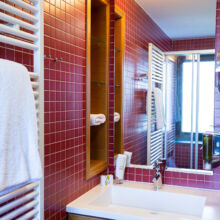 Modernes Badezimmer mit rot gefliesten Wänden, einem weißen Waschbecken mit Holzschrank, einem großen Spiegel, einem Wandregal mit Handtuchrollen, einem weißen Handtuch auf einem beheizten Handtuchhalter und natürlichem Licht durch ein Fenster.