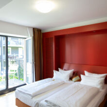Ein modernes Hotelzimmer verfügt über zwei Einzelbetten mit weißer Bettwäsche, eine auffällige rote Akzentwand und eine Balkontür mit Blick ins Grüne und auf die benachbarten Gebäude. Natürliches Licht durchflutet den aufgeräumten Raum.