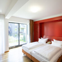 Ein modernes Hotelzimmer mit zwei sauber gemachten Einzelbetten, einer roten Akzentwand, einem großen Fenster mit durchsichtigen Vorhängen, einem kleinen Schreibtisch und einem Blick auf einen grünen Innenhof.