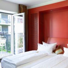 Ein modernes Hotelzimmer mit einem Doppelbett, weißem Bettzeug und einer orangefarbenen Akzentwand. Glastüren führen zu einem kleinen Balkon mit einem Tisch und Stühlen, von dem aus man auf andere Balkone des Gebäudes blickt. Das Zimmer ist von natürlichem Licht durchflutet.