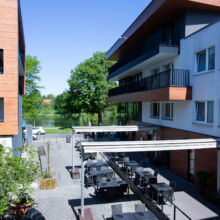 Ein moderner Apartmentkomplex mit Sitzgelegenheiten im Freien auf einem Innenhof zwischen zwei Gebäuden, mit Tischen und Stühlen unter einer Pergola, mit Blick auf einen See und Bäume an einem sonnigen Tag.