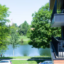Blick von einem Balkon mit Stühlen auf einen Park mit üppigen grünen Bäumen und einem ruhigen Fluss. Ein paar Leute gehen auf einem Weg spazieren, und Autos parken am Straßenrand unter blauem Himmel.
