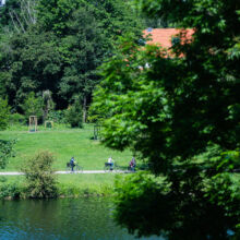 Drei Personen fahren mit ihren Fahrrädern auf einem Weg neben einem Fluss, umgeben von üppigen grünen Bäumen und Gras, wobei die Häuser im Hintergrund teilweise sichtbar sind.