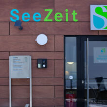 Gebäudeeingang mit einem Schild mit der Aufschrift SeeZeit in blauen und grünen Buchstaben, einem Licht, einer Sicherheitstastatur und einer Tür mit einem Herzlich Willkommen-Schild in Deutsch und Englisch.